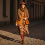 Kobieta w jesiennej stylizacji idzie po brukowanej ulicy wśród opadających liści